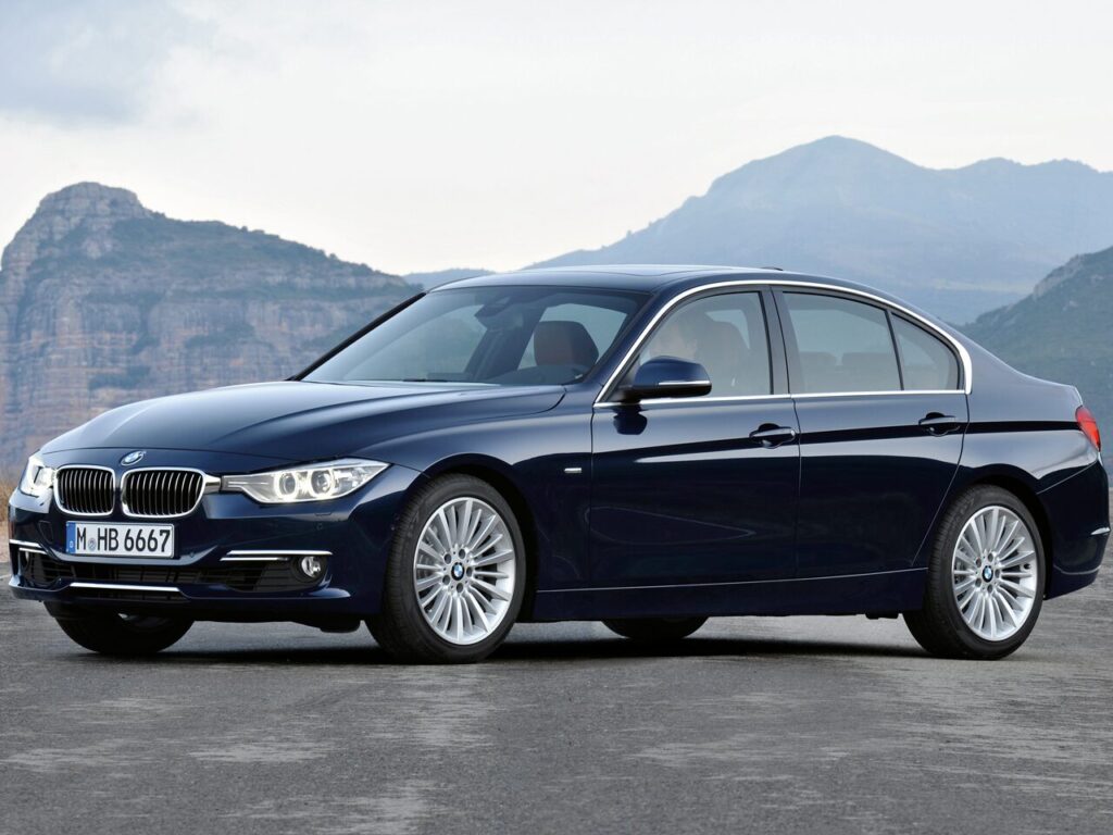 Front three-quarter view of a dark blue BMW sedan parked on a paved surface with mountains in the background.