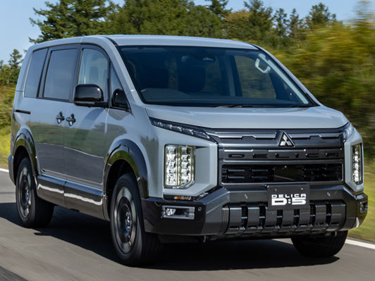 Silver Mitsubishi Delica D:5 minivan driving on a open road with trees in the background; front grille and vertical LED headlights visible.