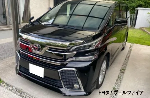 Front view of a black Toyota Vellfire minivan parked in a driveway beside a house, showing chrome grille and sleek headlights.