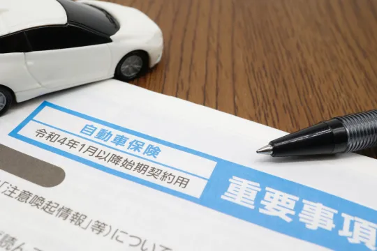 CG自動車保険 | 車買取のソコカラ White toy car sits on a wooden desk beside a Japanese auto insurance form with a blue header and a black pen nearby.