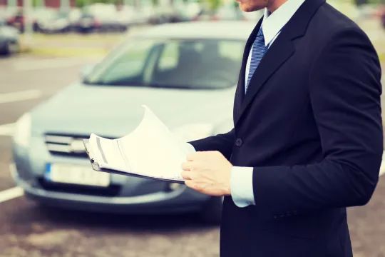 CG車査定 | 車買取のソコカラ Man in a dark suit and tie holds scattered documents in front of a parked car outside the frame edge shows the vehicle behind him.
