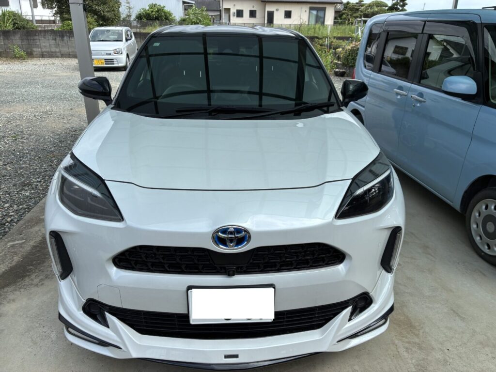ヤリスクロス ハイブリッド 2023年式 | 車買取のソコカラ Front view of a white Toyota SUV parked in a lot, with a blue car beside it and a small white car in the background under a covered area