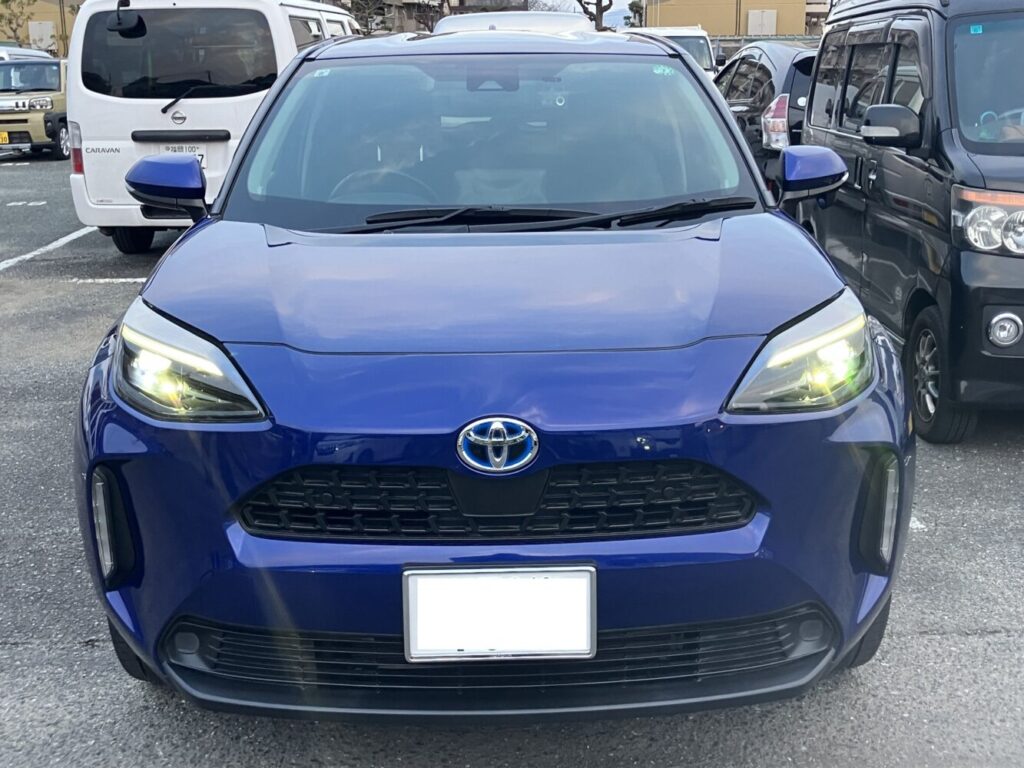 ヤリスクロス ハイブリッド2021 | 車買取のソコカラ Front view of a blue Toyota SUV with headlights on, parked in a crowded parking lot.
