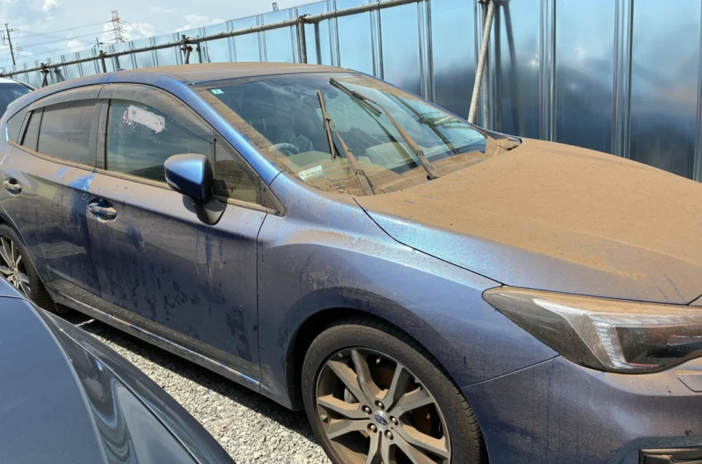 水没車 | 車買取のソコカラ Blue sedan parked in a lot, covered in a thick layer of dust and sand with windshield wipers resting on the windshield.
