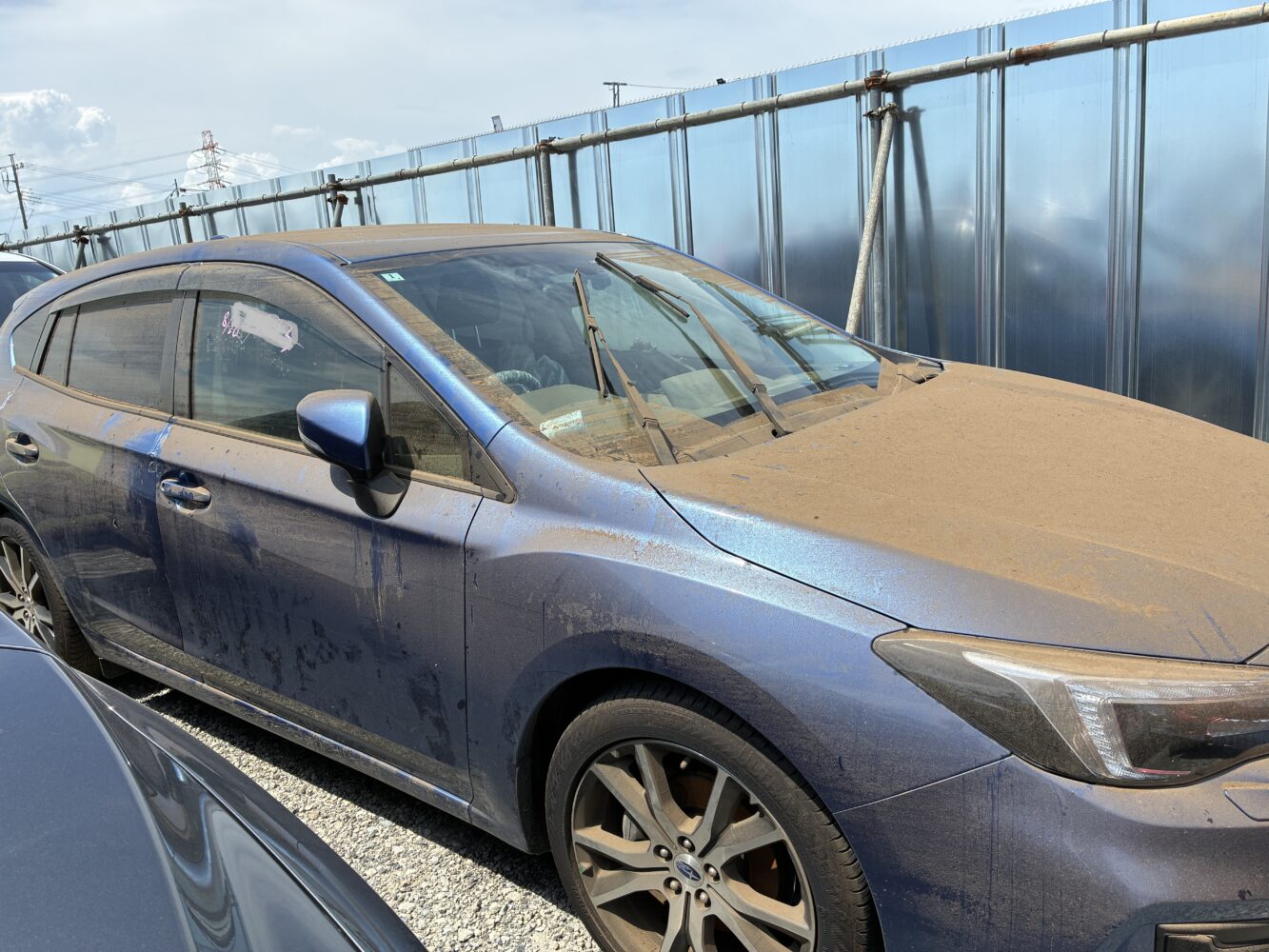 Dusty blue sedan parked in a lot beside a metal-framed fence on a sunny day, with windshield wipers raised and mud/dust on the surface.