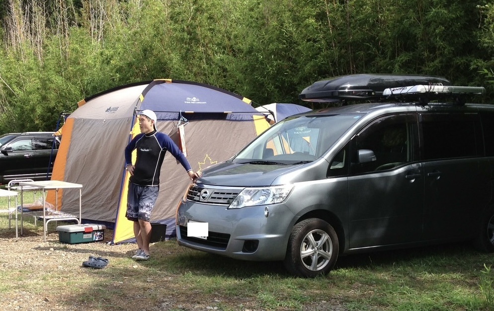 22_フォレスター | 車買取のソコカラ Man stands beside a gray van with a roof rack at a forest campsite, near a large blue-yellow tent.