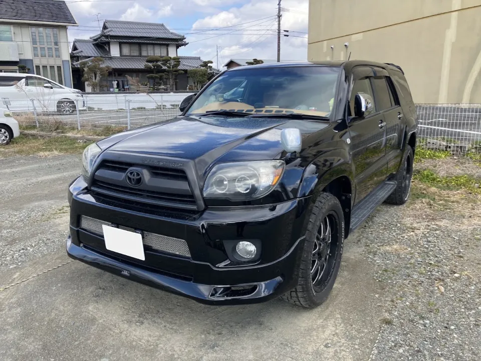 ハイラックス2006年式買取事例 | 車買取のソコカラ Black Toyota SUV parked on a gravel lot; front three-quarter view with houses and a fence in the background.