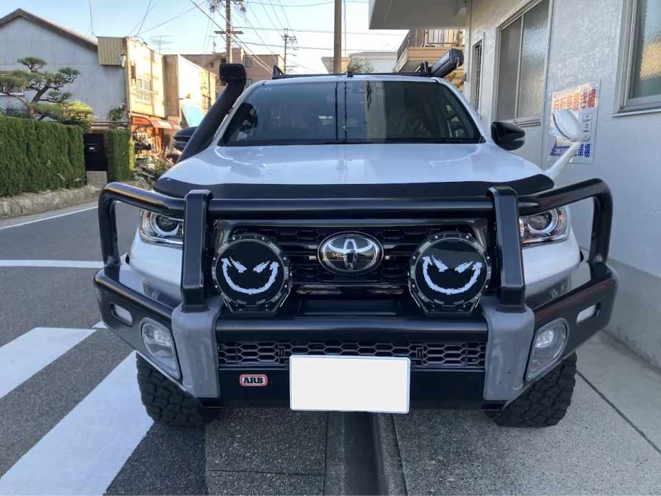 ハイラックス2019年式買取事例 | 車買取のソコカラ Front view of a white Toyota SUV with a heavy-duty bull bar, two round off-road lights, and a snorkel on a street.