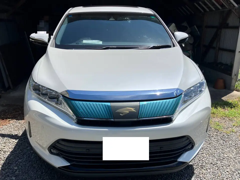 ハリアー買取事例_ハイブリッド | 車買取のソコカラ Front view of a white SUV parked on gravel, with a teal-accented grille and a front license plate area blanked out for privacy